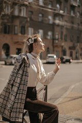 Young happy girl in pearl necklace enjoys sunny spring day. Pretty blonde woman in brown pants, white blouse and tweed jacket poses in city.