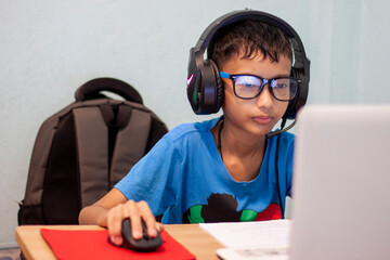 Asian boy wearing headphones and light filtering glasses is using laptops computers to study online at home.