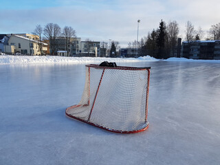 Outdoor ice hockey field, a goal, gloves and a stick at morning with an apartment building on the background.