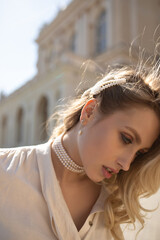 Portrait of charming blonde girl in pearl necklace and white blouse poses outside. Pretty woman...