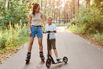 Family walk and rest in the summer day, mom riding roller skates and her son skates on a scooter in...