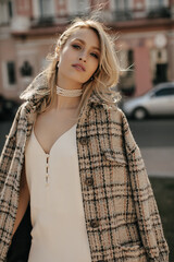 Stylish elegant blonde woman in white silk dress, checkered tweed coat and pearl necklace walking...