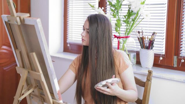 A young caucasian girl with long brown hair painting inside a studio. Focus on the girl. Bright windows and white flowers in the background.