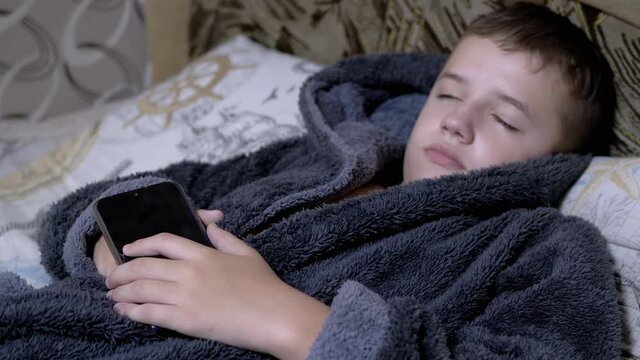 Adorable Child in Home Gray Dressing Gown Falls Asleep with Smartphone in Hands. A happy boy holds a mobile phone in his hands. A teenager is dependent on modern gadgets and tablets. Zoom. Close up.