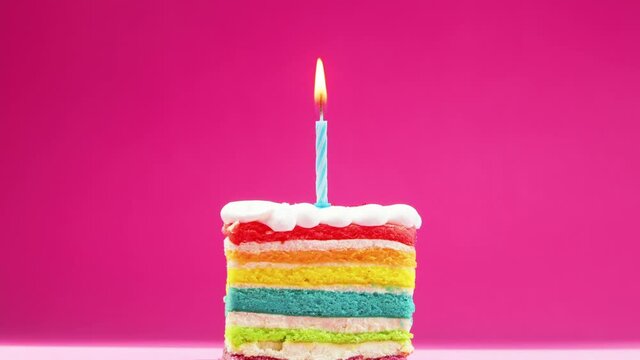 A slice of birthday cake with a bright yellow burning candle on a pastel pink background is blown out. A festive candle is burning on a rainbow piece of cake, which dies out and slowly smolders.