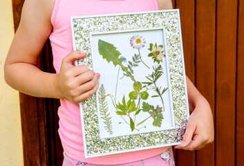Child showing handmade dried pressed real flowers plants in picture frame. Arts and crafts concept....