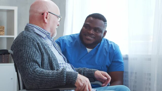 African-American caregiver and old disabled man in a wheelchair. Professional nurse and handicapped patient in a nursing home. Assistance, rehabilitation and health care.