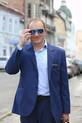 Portrait of young confident Caucasian man in blue business suit looking forward while holding sunglasses with hand. The background is the bustling business center of the city. Europe. Work, business
