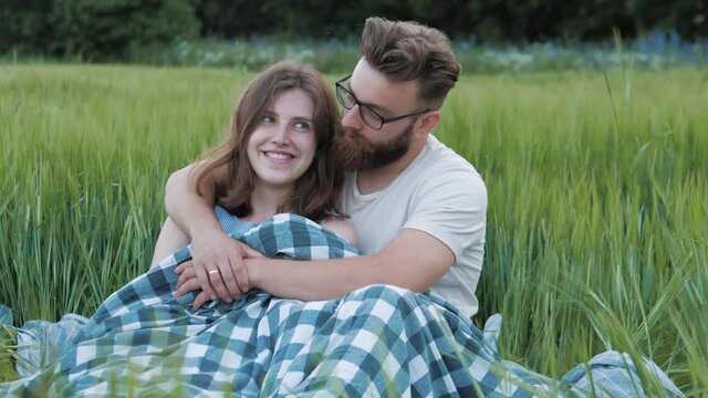Bearded man hugging a woman, having a date on a green field. Boyfriend and girlfriend sitting under blanket enjoying moments together, true love, young family