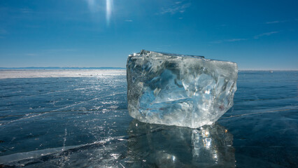 A transparent ice crystal lies on the smooth blue surface of a frozen lake. Glare of the sun on the...