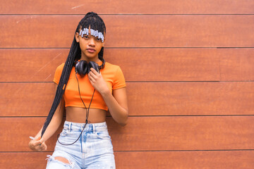 Lifestyle with a young trap dancer in the city. Black grind girl of African ethnic group with orange t-shirt and cowboy pants, with headphones on a wooden background