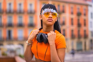 Street Style of a young trap dancer with braids. She pose from the black grinding girl of African ethnic group with t-shirt, orange sunglasses and cowboy pants sitting. With orange houses background