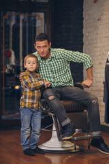 Fototapeta na wymiar Young handsome father and his little stylish son at barbershop waiting for barber