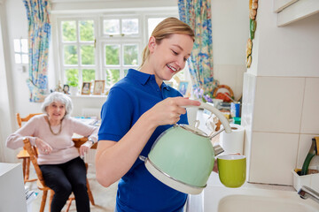 Female Home Help Making Cup Of Tea In Kitchen Whilst Chatting With Senior Woman