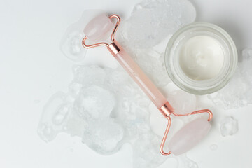 Concept of cooling skin care for summer. Closeup of a jar of moisturizer and a rose quartz roller surrounded by ice.