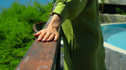 European girl stands in the summer on a balcony holding her hand to a wooden railing in a green dress