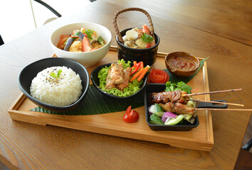 asian bento party set with seafood soup, deep fried snack, chicken satay and peanut spicy sauce, stir fried vegetables and steam white rice on wood board tray asian halal menu