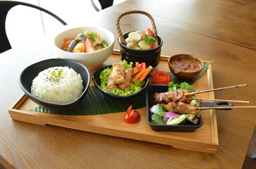 asian bento party set with seafood soup, deep fried snack, chicken satay and peanut spicy sauce, stir fried vegetables and steam white rice on wood board tray asian halal menu