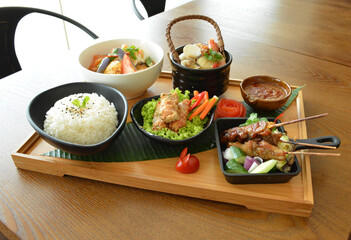 asian bento party set with seafood soup, deep fried snack, chicken satay and peanut spicy sauce, stir fried vegetables and steam white rice on wood board tray asian halal menu