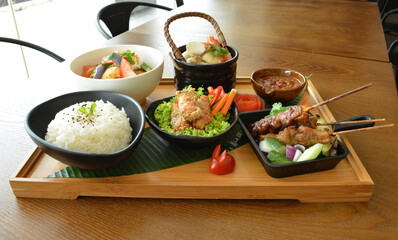 asian bento party set with seafood soup, deep fried snack, chicken satay and peanut spicy sauce, stir fried vegetables and steam white rice on wood board tray asian halal menu