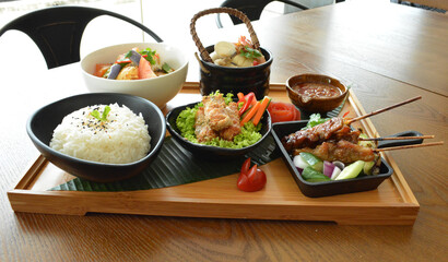 asian bento party set with seafood soup, deep fried snack, chicken satay and peanut spicy sauce, stir fried vegetables and steam white rice on wood board tray asian halal menu