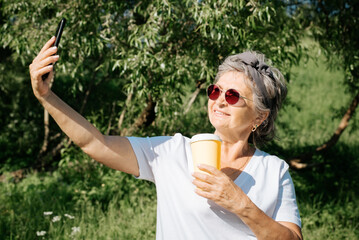 Fototapeta premium Modern smiling mature woman in red glasses and cup of coffee taking a selfie on mobile phone while standing in park, outdoors. Trendy beautiful senior woman using smartphone outside, summer lifestyle