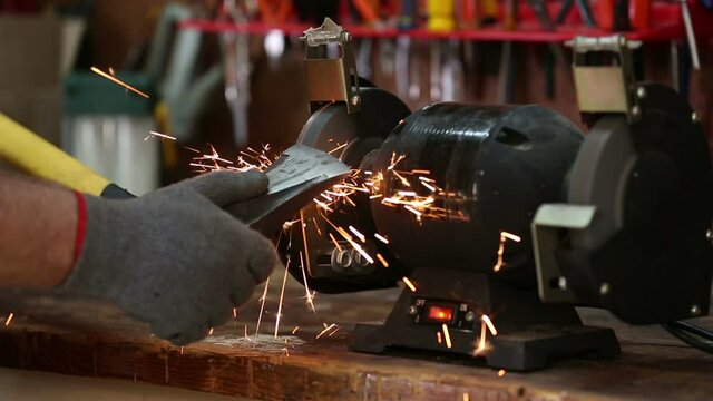 Sharpening an axe wearing gloves on a grinder with sparks flying on workbench