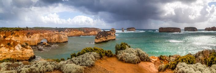 A huge panorama of rocky reddish cliffs and small islands in the southern ocean. Victoria. Great Ocean Road. A line of protruding rocky islands on the horizon. The sky is covered with dark clouds.