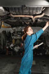 A girl auto mechanic checks the chassis of a car and bridges in a car repair shop or garage. Car repair and locksmith with glasses and special suit at work. Diagnostics of the car.