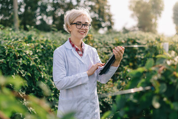 Agronomist at work. Portrait of young woman agronomist at plantation. Worker with tablet smiles and looks into camera. - obrazy, fototapety, plakaty