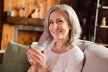 Photo of shiny sweet lady pensioner wear white t-shirt sitting sofa smiling drinking coffee indoors...