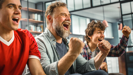 At Home Three Joyful Soccer Fans Sitting on a Couch Watch Game on TV, Celebrate Victory when Sports...