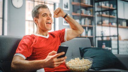 Charismatic Young Adult Man Sitting on a Couch Watches Game on TV, uses Smartphone App for Score,...