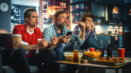 Night at Home: Three Soccer Fans Sitting on a Couch Watch Game on TV, Use Smartphone App to Online Bet, Celebrate Victory when Sports Team Wins. Friends Cheer Eat Snacks, Watch Football Play.