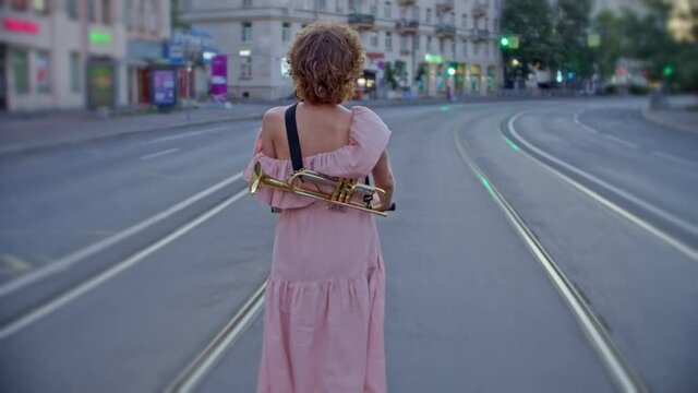 view from the back of a loose woman in a summer dress with a musical instrument rides in the middle of the empty road of the extinct city along the tram tracks, the dress develops. the image of a free