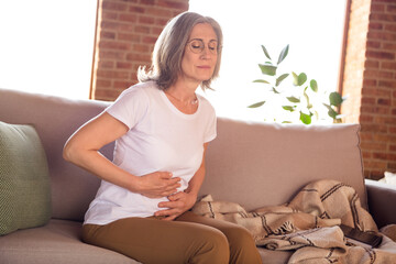 Photo of unhappy upset lady pensioner wear white t-shirt sitting sofa arms stomach having pain indoors house flat