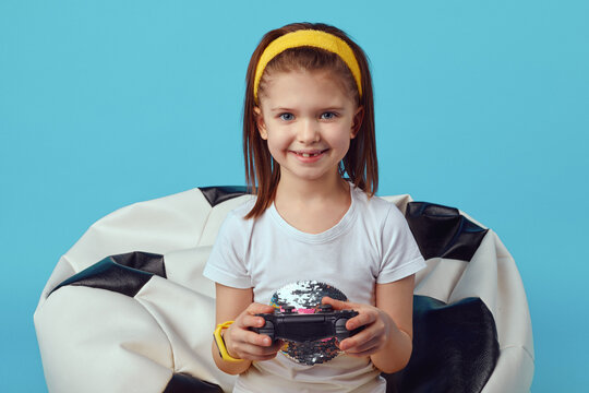 Full length photo of happy little girl sitting on bean bag chair, holding joystick and playing video games, wearing sport outfit, isolated over blue