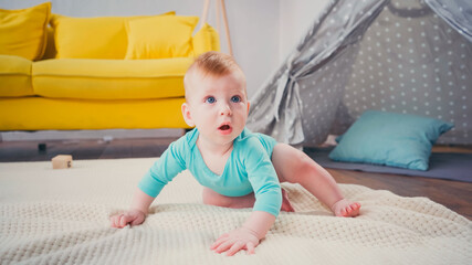 surprised boy in romper crawling on blanket near blurred tipi