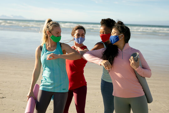 Group of diverse female friends wearing face mask practicing yoga,standing and at the beach