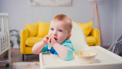 infant boy with blue eyes sitting in feeding chair and sucking spoon near bowl