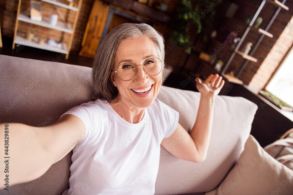 Sticker Self-portrait of attractive cheerful grey-haired woman sitting on divan welcoming you at loft industrial home house flat indoors