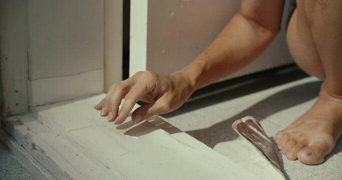 Close up of young woman hands removes dry paint in doorstep at sunny morning. European girl preparing to paint the door in her house. Hand close-up doing home renovation.