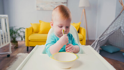 infant boy sitting in feeding chair and holding spoon near bowl