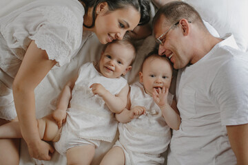Obraz premium Top view of charming family portrait with baby twins. Smiled parents with firsborns in love dressed in white clothing, laying at bedroom, weekend, family vacation. Concept of happy healthy parenthood