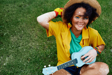 Playful African American woman with blue ukulele having fun while sitting on green lawn wearing...