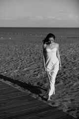 black and white photo girl in white long in a dress and short hair walks at sunset on the sand near the sea