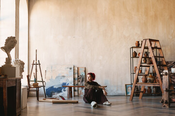 Young woman artist sitting on a floor of an art studio - obrazy, fototapety, plakaty