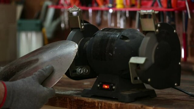 Sharpening a shovel wearing gloves on a bench grinder with sparks flying on workbench