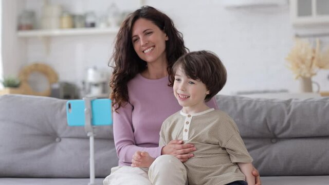 Family Video Call: Joyful Mom And Kid Of Five Waving Hands To Smartphone On Tripod Say Hi To Grandparents Meeting Relatives Online On Videocall During Lockdown Quarantine Happy Smiling Sit On Couch