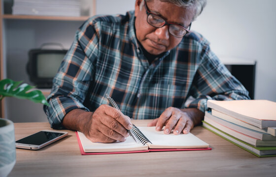 Elderly Asian man farmer with pen writing on notebook on his desk at home.